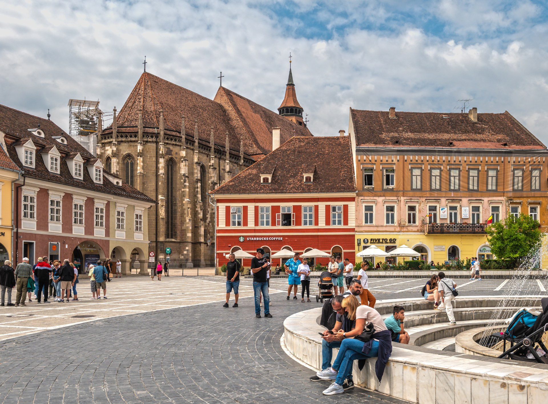 Mladí lidé odpočívají u kašny na Náměstí Rady v Brašově, v pozadí Černý kostel